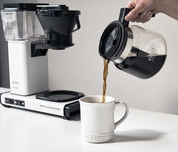 A picture of some pouring a cup of coffee with a coffee maker in the back ground on a white counter top.